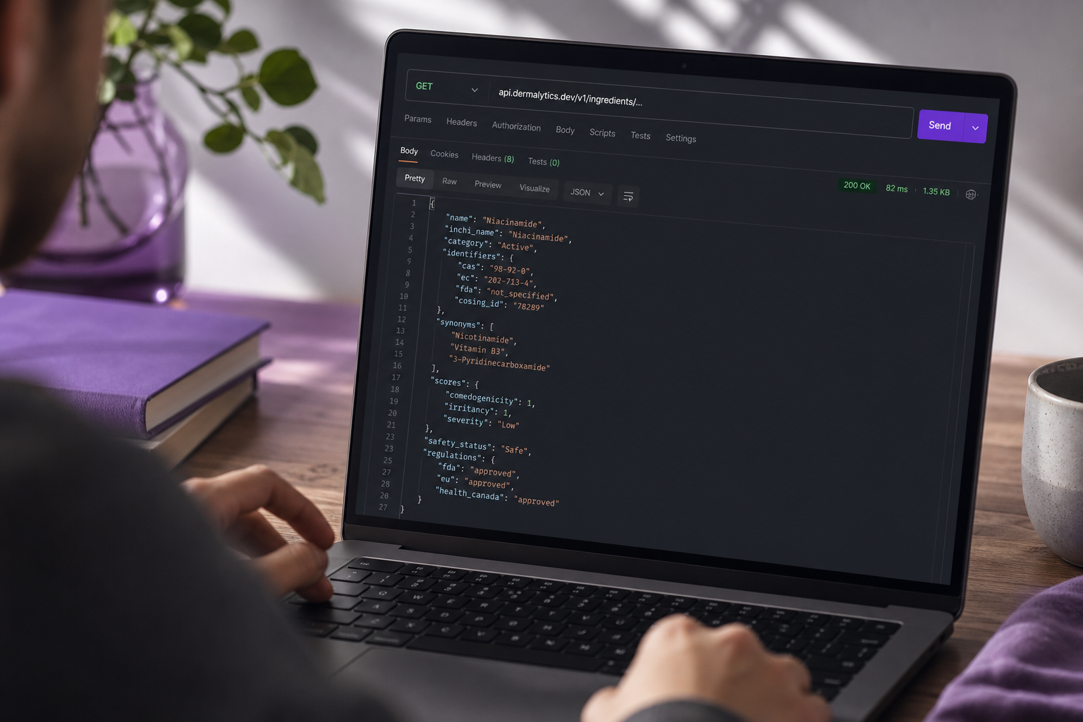 Close-up over-the-shoulder shot of a developer's screen showing a Postman or Insomnia API client window — the URL bar reads "api.dermalytics.dev/v1/ingredients/..." and a formatted JSON response is visible in the response pane. Soft natural