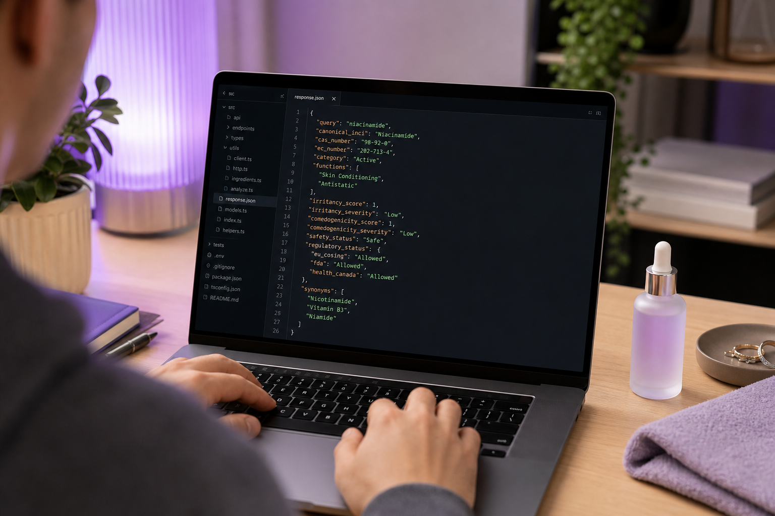 Over-the-shoulder developer workstation shot: laptop showing JSON response with fields like canonical_inci, cas_number, irritancy_score, comedogenicity_score, safety_status; a skincare bottle on the desk beside the keyboard. Warm office lighting, rea