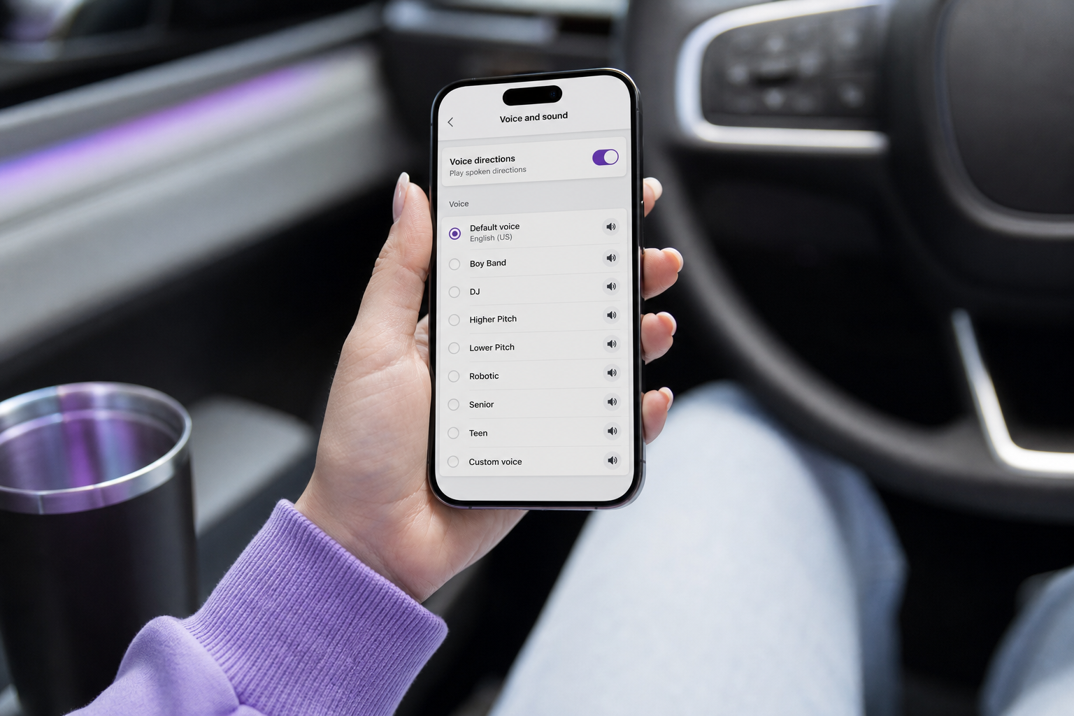 A smartphone held in one hand showing the Waze "Voice and sound" settings screen with a voice-selection list visible, car interior softly blurred in the background.